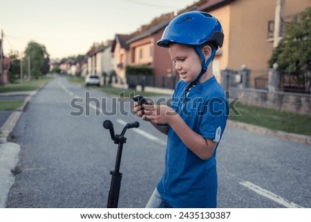 Photo of a little boy using a mobile phone while riding a push scooter outdoors.