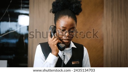 Portrait of busy successful woman working as receptionist in luxury hotel. Attractive African-American female receptionist receiving calls talking on phone. Friendly young hotel administrator. Royalty-Free Stock Photo #2435134703