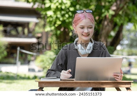 Asian female student studying online Happy woman looking at computer screen Surf the web or do video chat on webcam. Happy woman using laptop and wearing headphones sitting in the park.