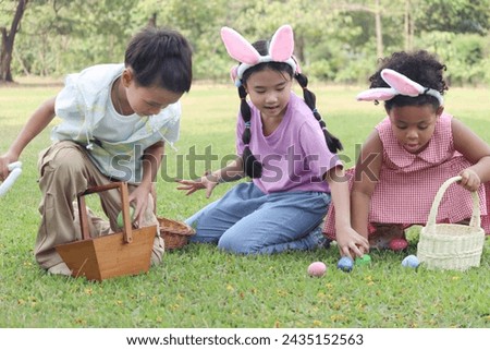 Three little children with bunny ears have fun in park. Asian girl and boy with curly hair African child friend hunting Easter eggs in green garden. Kids celebrating Easter spring holiday at outdoor.