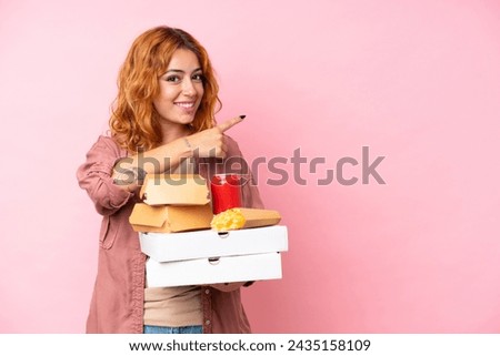 Young caucasian woman holding fast food isolated on pink background pointing back