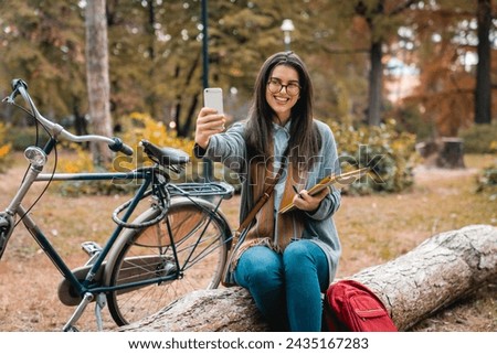 Attractive young college girl sitting on the tree trunk in public park and taking selfies while waiting for her friend to appear.