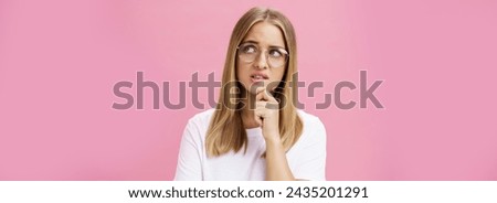 Studio shot of insecure smart nerdy woman in glasses and white t-shirt standing troubled and worried touching chin looking bothered at upper right corner hesitating, thinking against pink wall.