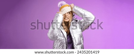 Awkward embarrassed cute blond girl worried pulling hat face hiding from ex-boyfriend pretend not see him standing anxiously clenching teeth peeking one eye left nervous, purple background.