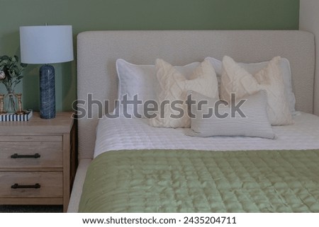 Cozy bed with green comforter or duvet with many white pillows, upholstered headboard and wood side table Royalty-Free Stock Photo #2435204711