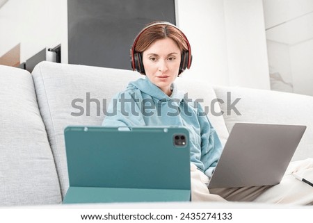 A young girl sits on the sofa and studies hard using many gadgets. Technology and education concept Royalty-Free Stock Photo #2435274133