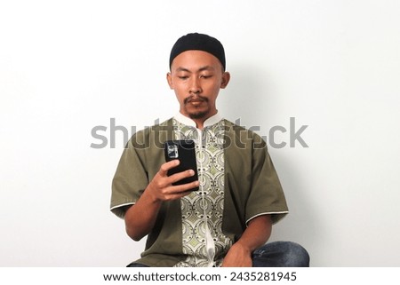 Bored Indonesian Muslim man in koko and peci scrolls through his phone, seeking distraction while fasting during Ramadan, Waiting for Iftar. Isolated on a white background