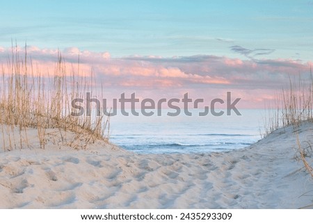 Sandy path of footprints leading through the dunes and grasses to the beach and the Atlantic Ocean under a pastel sky of pink and blue at sunset on the Outer Banks of North Carolina. Royalty-Free Stock Photo #2435293309