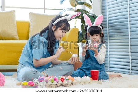 Asian cute little children girl wearing funny bunny ears headbands and young pretty mother smile decorating painting eggs while sitting on floor in living room, Family preparing for Easter holiday.