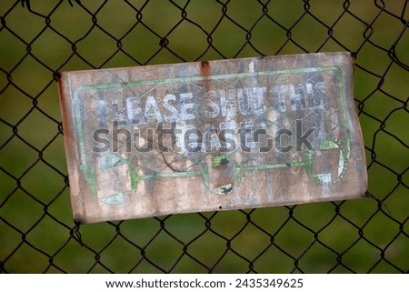 View of an old faded sign on a sheet of tin attached to a rusty chain wire fence asking to please shut the gate