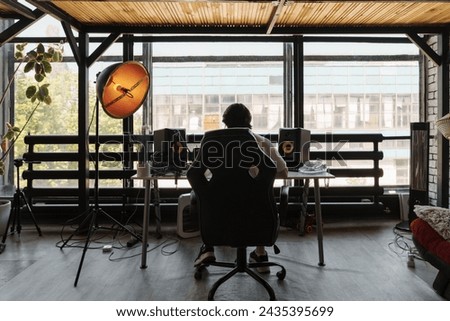 Back view of unrecognizable male musician sitting at table with loudspeaker enclosure near panoramic window and composing music Royalty-Free Stock Photo #2435395699