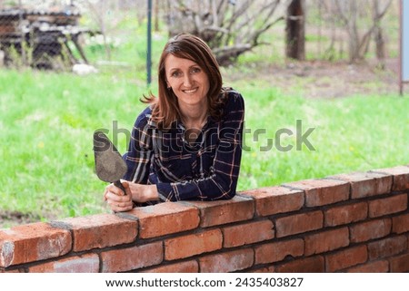 a young female bricklayer with a trowel in her hands is smiling, leaning against a brick wall