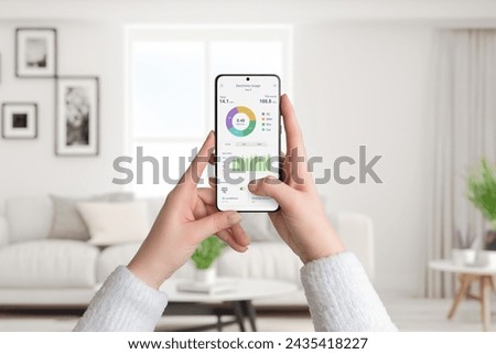 Woman holds smartphone with electricity usage app, monitoring and analyzing consumption for smart home. Living room background. Concept of energy efficiency