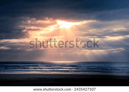 Sunset over Muriwai beach. Sun beams breaking through heavy clouds as waves splashing into wet sand. Auckland, New Zealand. Royalty-Free Stock Photo #2435427107