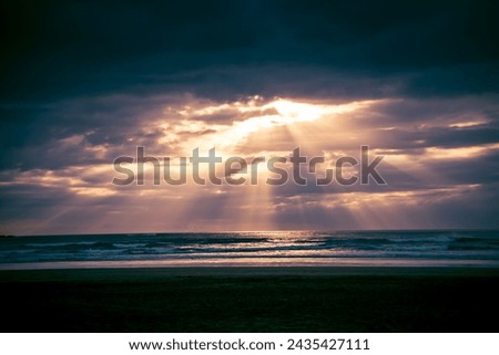 Sunset over Muriwai beach. Sun beams breaking through heavy clouds as waves splashing into wet sand. Auckland, New Zealand. Royalty-Free Stock Photo #2435427111