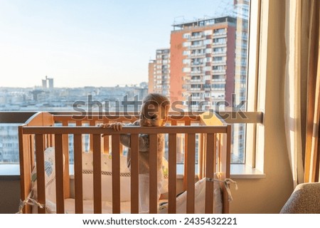 the baby in the crib stands and is bored looking inside the room, not paying attention to the landscape outside the window