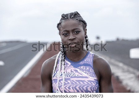African American female runner with braids looking at camera while standing on blurred background of road during outdoor training