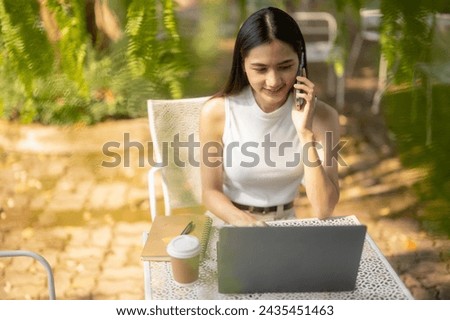 A young woman talks on a cell phone while using a laptop in an outdoor cafe setting with coffee. Talk and discuss via smartphone.