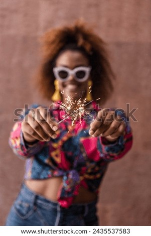 Blurred chappy ethnic female in trendy clothes crossing sparklers while having fun against brown background Royalty-Free Stock Photo #2435537583