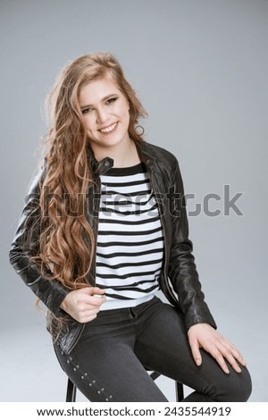 woman with long brown hair is sitting on chair and smiling. She is wearing black and white striped shirt and leather jacket. Concept confidence and style, as woman is posing for photo