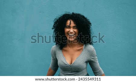 Joyful South American young woman laughing and smiling. authentic burst of laughter of a black adult girl in 20s on urban environment, teal backdrop Royalty-Free Stock Photo #2435549311