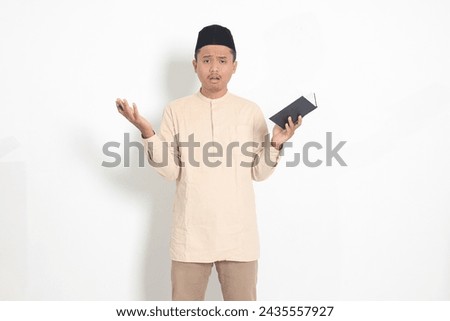 Portrait of confused Asian muslim man in koko shirt with peci difficulty understanding the contents of the book, reading a textbook. Isolated image on white background Royalty-Free Stock Photo #2435557927