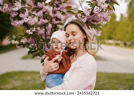 Mom holding his baby in the arms near the cherry blossom in the park. Blonde woman holding small child near sakura. 