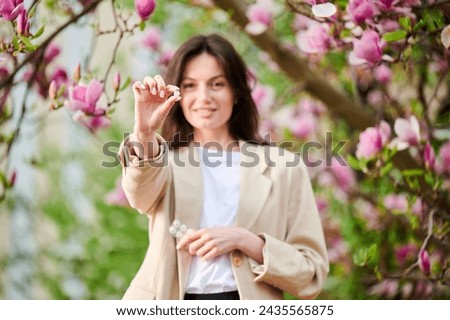 Woman allergic suffering from seasonal allergy at spring, posing in blossoming garden. Focus on woman's hand showing allergy drugs, holding pack of pills. Antihistamine medication concept
