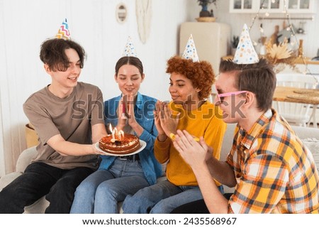 Make a wish. Woman wearing party cap blowing out burning candles on birthday cake. Happy Birthday party. Group of friends wishes girl happy birthday. People celebrating birthday with party at home