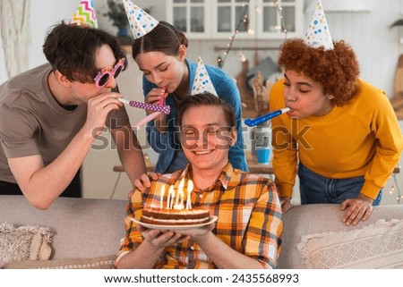 Make a wish. Man wearing party cap blowing out burning candles on birthday cake. Happy Birthday party. Group of friends wishes guy happy birthday. People celebrating birthday with party at home