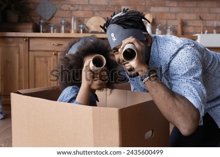 Cute African girl sit inside carton box play pirates with father, family play astronomers, holding paper tubes watching into distance at camera pretend to be explorers. Funny playtime at home concept Royalty-Free Stock Photo #2435600499