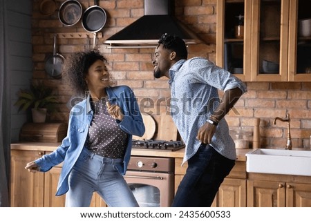 Lively millennial African hipster couple dancing in kitchen at home. Vivacious wife and husband enjoy weekend, listen favourite music moving indoor looking overjoyed, homeowner family leisure concept Royalty-Free Stock Photo #2435600533
