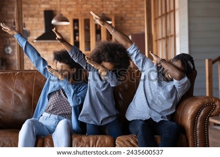 Happy African couple and little 6s daughter sit on sofa showing dab gesture, raise their hands up hides faces having fun fooling at home, do popular internet meme pose, perform triumph dance concept