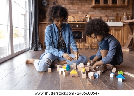African mom and daughter play wooden bricks at home, sit on warm floor in domestic kitchen, build use color cubes, enjoy playtime and carefree weekend. Developmental games and growth of child concept Royalty-Free Stock Photo #2435600547
