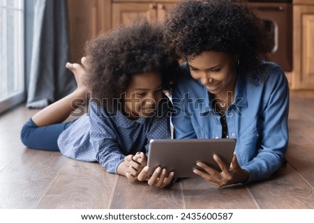 African woman her little 6s daughter lying on warm floor in kitchen, spend leisure at home use digital tablet, enjoy new amusing apps, watch internet videos, users of e-shopping and e-commerce concept Royalty-Free Stock Photo #2435600587