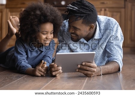 African ethnicity father little 6s cute daughter spend time at home lying on warm floor in kitchen using digital tablet, watch videos, play online games. Wireless modern tech usage, have fun concept