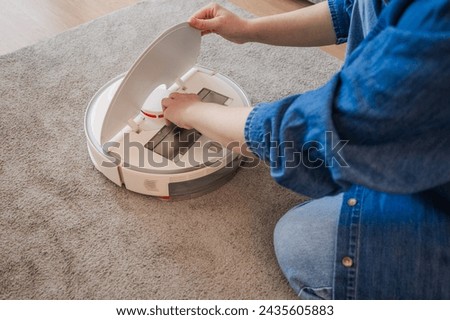 Young caucasian woman opening an automatic vacuum cleaner to clean the floor at home in living room, replace the filter. smart devices, modern technologies concept Royalty-Free Stock Photo #2435605883