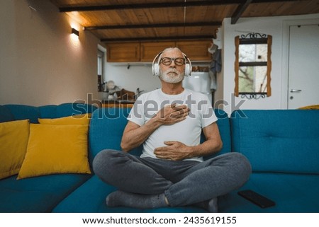 One man senior caucasian male eyes closed for guided training yoga or meditation while sitting at home with headphones self-care practice real people well-being inner peace and balance concept Royalty-Free Stock Photo #2435619155