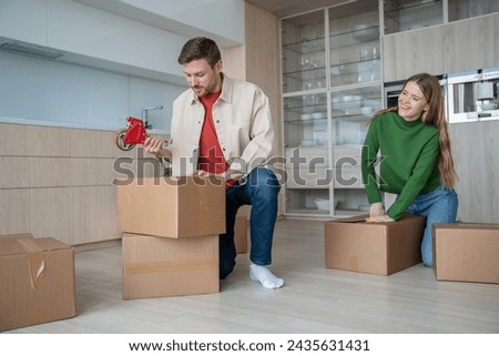 Couple energetically emptying rented apartment, putting belongings in cardboard boxes. Guy and girl carefully packing in preparation for long-awaited move. New chapter young family life your own home. Royalty-Free Stock Photo #2435631431