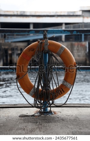 Exterior photo visual view of an orange plastic lifebuoy with a rope attachde hooked to a etal gate fence at the harbour near the bak platform for safety  for people who fall in water and swim help
