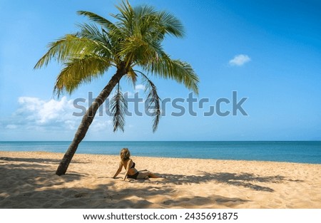 A young woman sits on the sand in the shade of a palm tree on a tropical island