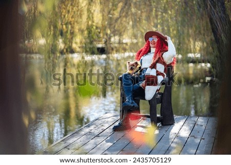 Autumn lake woman red hair dog. She sits by a pond on a wooden pier in autumn and admires nature, holding a Yorkshire terrier in her hands. The concept of tourism, weekends outside the city.