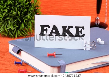 FAKE word written on a business card standing with a clip on a diary, a notebook on an orange background