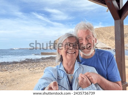 Cheerful senior smiling caucasian couple hugging standing outdoors together at the beach takes photos with selfie stick enjoying vacation and retirement