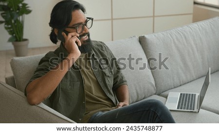 Muslim Arabian guy freelancer sit on couch with laptop talk mobile phone at home smiling Indian man businessman in eyeglasses speak smartphone answer call carefree converse cell business discussion Royalty-Free Stock Photo #2435748877