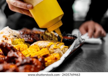 A close-up of someone drizzling mustard onto grilled corn, served alongside BBQ ribs and coleslaw on a tray. The ribs are well-cooked, and the background appears to be a kitchen or restaurant