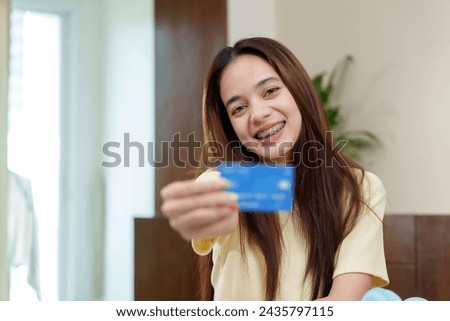 Cheerful Asian lady displays a blue credit card, ready for online shopping, comfortable workspace. Smiling woman with dental braces presents a card, set for an online purchase, inviting home setting