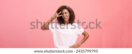 Not afraid express myself. Joyful charismatic african american woman in t-shirt with afro haircut showing tongue playfully and daring making peace sign over eye and winking posing over pink background
