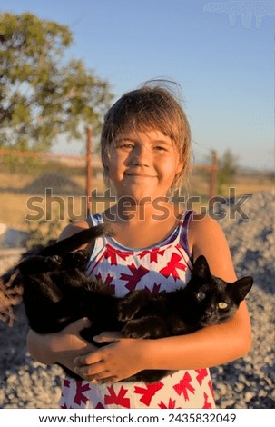 A black cat in the arms of a child outdoors. The cat looks helplessly and seems to want to escape.