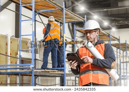 Man builder with electronic tablet. Engineer in construction uniform. Foreman near scaffolding. Builder guy in industrial building being renovated. Construction of factory. Man builder doing design
